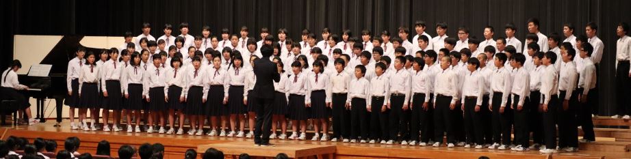 ステージ上でピアノを伴奏している生徒の横に複数の児童が指揮者に合わせて合唱している様子を観客が見ている写真