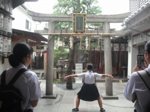鳥居の前で1人の女子生徒が大きく腕と足を広げその様子を2人の女子生徒が見ている写真