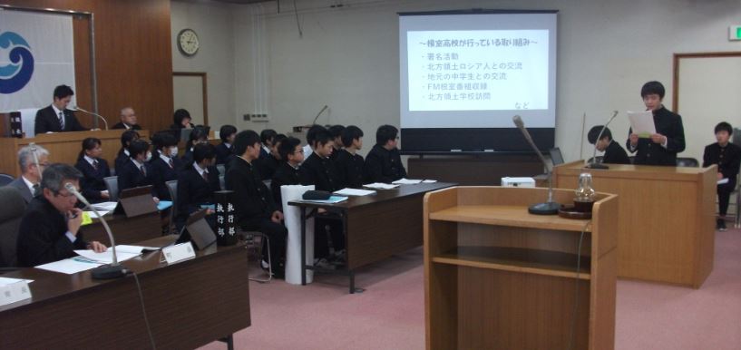 役場議場で1人の男子生徒が資料を手に持ちマイクに向かって話している様子を複数の人が見ている写真