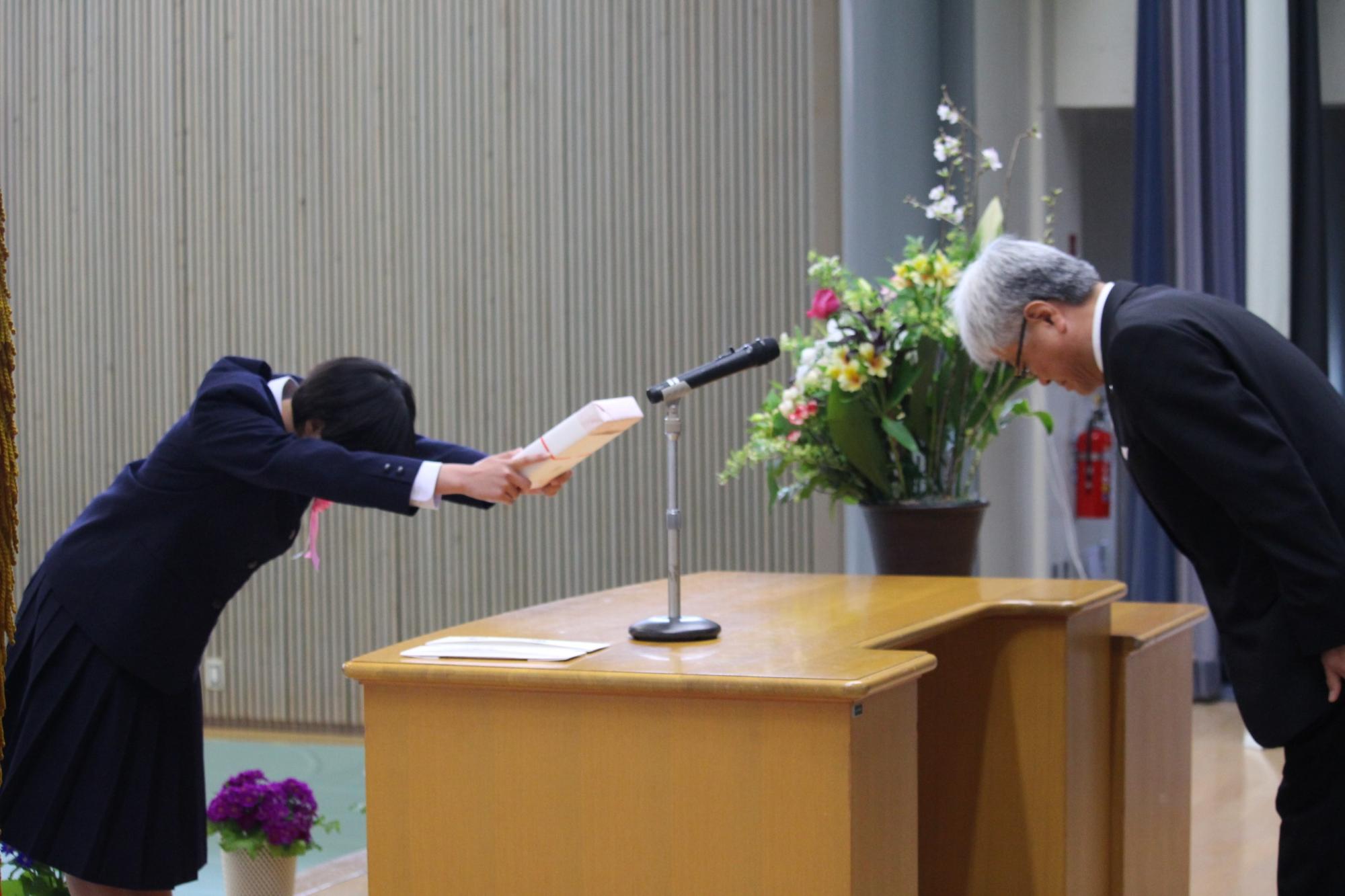 新入生がステージで年配の男性と向き合って物を受け取りお辞儀をしている写真