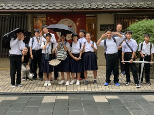 瓦屋根の建物の前で生徒達が並び、傘を差したりポーズをとったりして記念撮影している写真