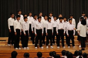 ステージ上で男子生徒達が並び女性指揮者の前で合唱している写真