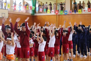 体育館で大勢の生徒と児童達が両手を挙げている写真