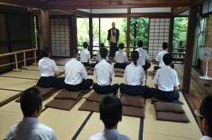 和室で複数の生徒が胡座をかいて座布団に座り僧侶の話を聞いている写真
