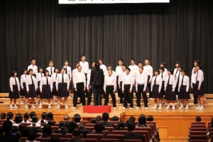 ステージ上に複数の生徒が肩幅に両足を広げ、指揮者を見ながら合唱している写真