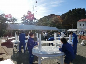 屋外で体操服姿の複数の生徒が丸テーブルを運んでいる写真