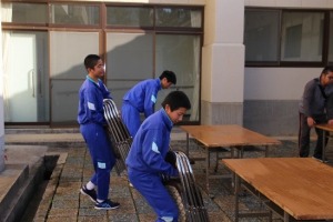 屋外で複数の生徒がパイプ椅子を幾つか手に持ち運んでいる写真