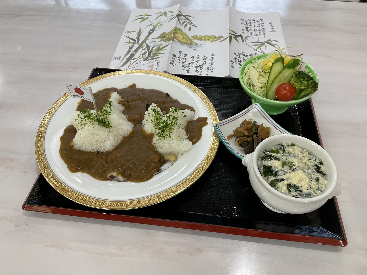 竹島の形をしたライスに日本国旗が立てられている竹島海鮮カレーの写真。お盆の上にはカレー、わかめスープ、サラダ、漬物がある。