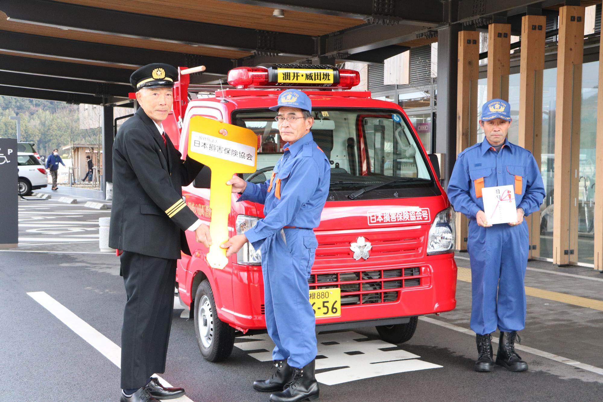 寄贈された消防車の前で制服を着た男性と作業着姿の男性が並んでレプリカキーと目録を持っている写真